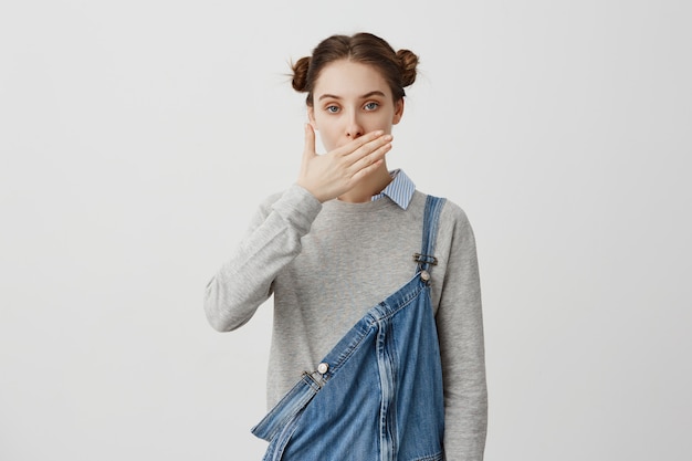 Morena mujer 30 años cubriendo la boca con la mano siendo tranquila. Mujer segura en denim casual que se niega a hablar permaneciendo en silencio. Gente, concepto de actitud