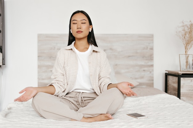 Morena mujer asiática en auriculares meditando en el dormitorio