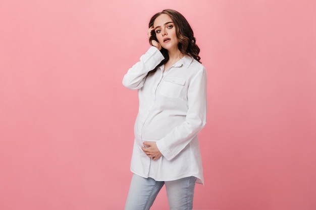 Morena mujer embarazada con camisa blanca mira a cámara. Chica joven alegre en jeans posa sobre fondo rosa aislado.