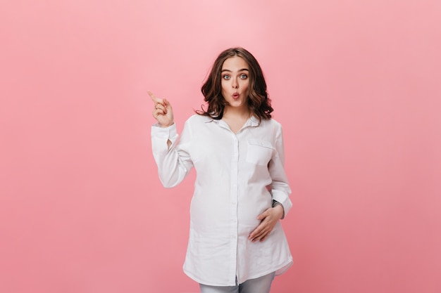 Morena de ojos azules mujer embarazada con camisa blanca y pantalones de mezclilla silba y señala el lugar para el texto sobre fondo rosa aislado.