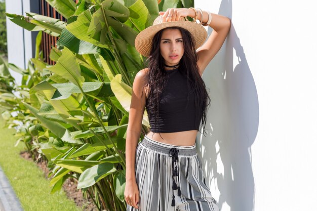 Morena sexy con cuerpo bronceado en sombrero de paja posando al aire libre. Tendencias de la moda de verano. Estado de ánimo tropical. Pulseras de madera.