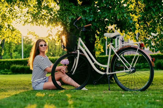 Morena se sienta en el césped verde con bicicleta y sostiene un ramo de flores en el parque.