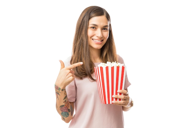 Morena sonriente mujer apuntando al cubo de palomitas de maíz sobre fondo blanco.