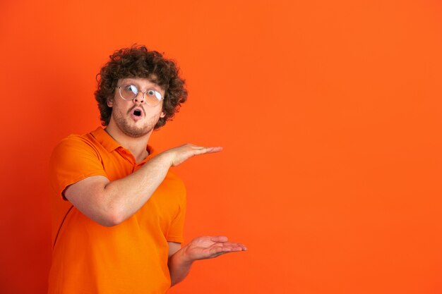 Mostrando algo grande. Retrato monocromático del hombre joven caucásico en la pared anaranjada. Hermoso modelo masculino rizado en estilo casual. Concepto de emociones humanas, expresión facial, ventas, publicidad.