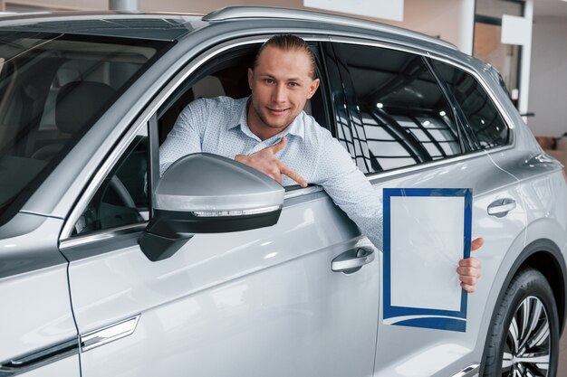 Mostrando con el dedo índice. Gerente sentado en coche blanco moderno con papel y documentos en manos