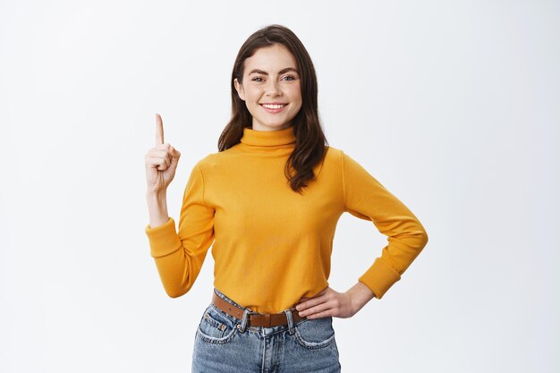 Mostrando las mejores ofertas. Guapa mujer morena sonriente apuntando con el dedo hacia arriba en blanco