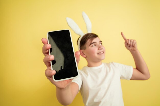 Mostrando pantalla en blanco, apuntando hacia arriba. Chico caucásico como un conejito de Pascua sobre fondo amarillo. Saludos felices de pascua.