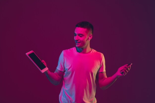 Mostrando pantalla en blanco. Retrato de hombre caucásico aislado en la pared rosa-violeta en luz de neón. Modelo masculino con dispositivos. Concepto de emociones humanas, expresión facial, ventas, publicidad. Copyspace.