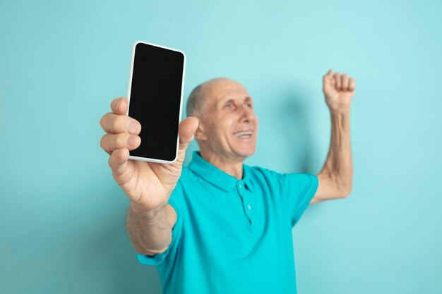 Mostrando la pantalla del teléfono en blanco. Retrato de hombre mayor caucásico en estudio azul.