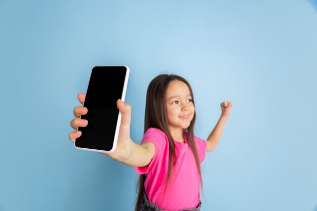 Mostrando la pantalla del teléfono. Retrato de niña caucásica en la pared azul.