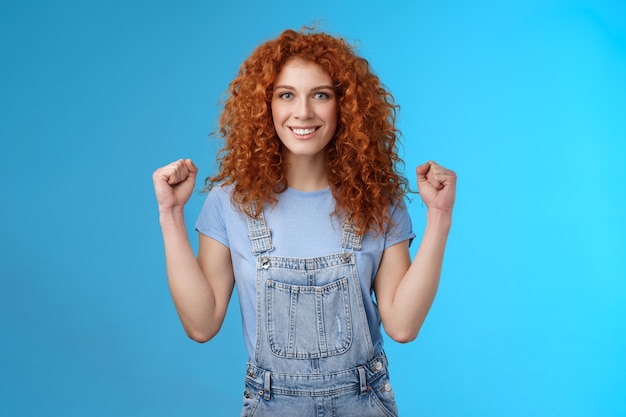 Motivada, decidida, segura de sí misma, atractiva pelirroja alegre chica aprieta los puños Mujer empoderada aseguró que todo salió bien día de verano afortunado sonriendo encantado fondo azul confiado.