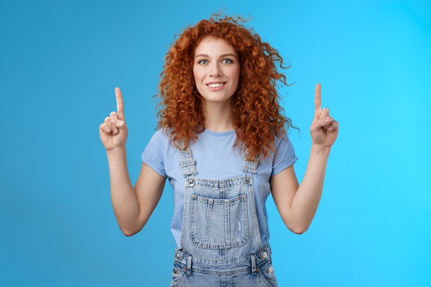 Motivado feliz alegre pelirroja tonta mujer rizada apuntando hacia arriba con el dedo índice sonriendo encantado impresionado emocionado mostrando promo impresionante discutir fondo azul de anuncio interesante.