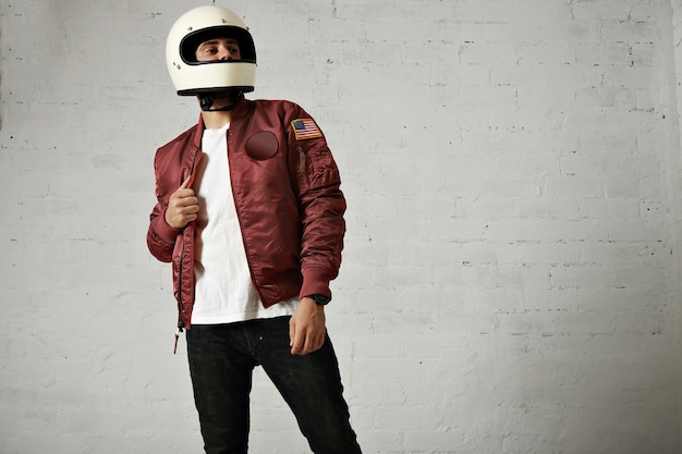 Motociclista de aspecto orgulloso en un casco blanco liso, chaqueta de bombardero de nailon burdeos, jeans y camiseta sobre fondo de pared blanca