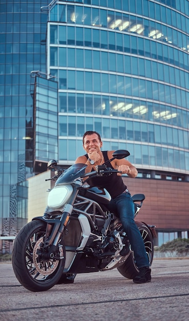 Un motociclista guapo y brutal vestido con una camiseta negra y jeans, con una sonrisa encantadora, sentado en una motocicleta deportiva, contra un rascacielos en el centro de la ciudad.