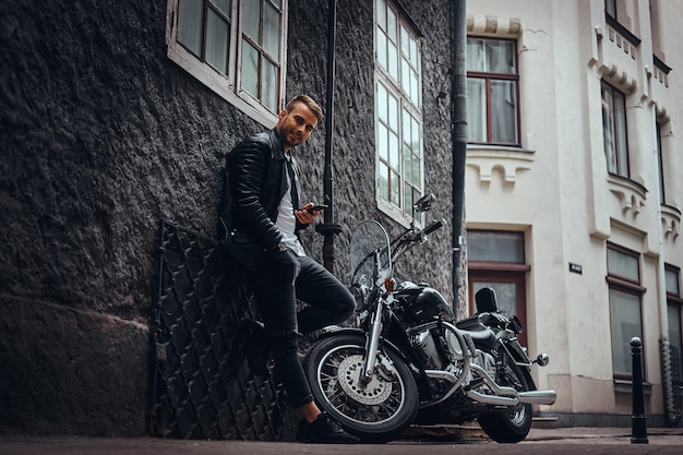 Un motociclista de moda sonriente vestido con una chaqueta de cuero negro y jeans sostiene un teléfono inteligente apoyado en una pared cerca de su motocicleta retro en una vieja calle de Europa.