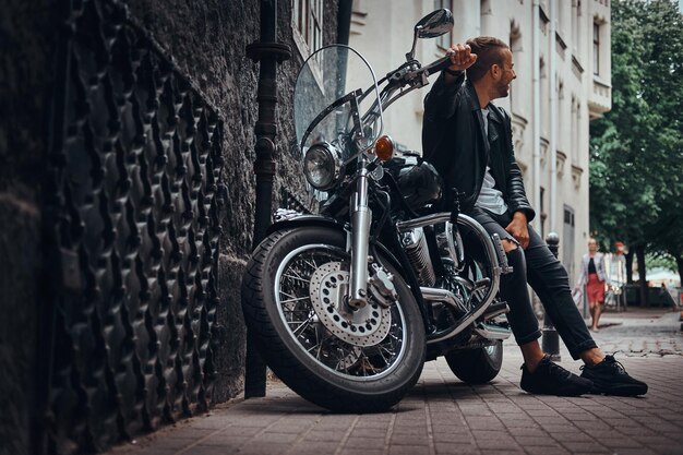 Motociclista de moda vestido con una chaqueta de cuero negro y jeans sentado en su motocicleta retro en la calle Old Europe.