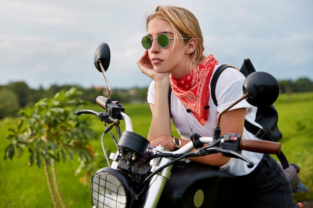 La motociclista reflexiva viste elegantes cortinas de verano, pañuelo y camiseta, lleva una mochila, se sienta en su moto rápida, cabalga por la naturaleza verde