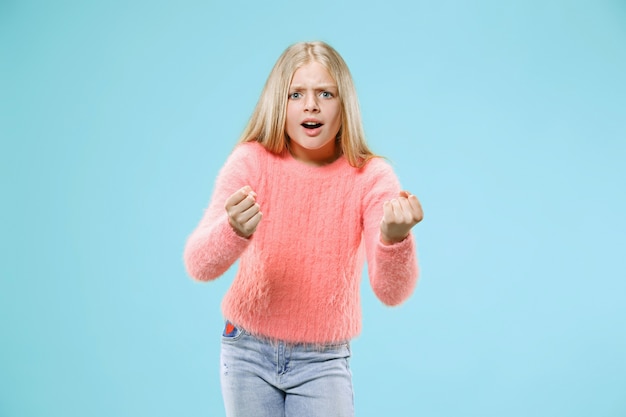 Muchacha adolescente enojada que se coloca en el fondo azul de moda del estudio. Retrato femenino de medio cuerpo. Las emociones humanas, el concepto de expresión facial.