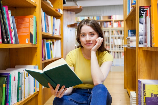 Muchacha adolescente con el libro que mira la cámara entre los estantes