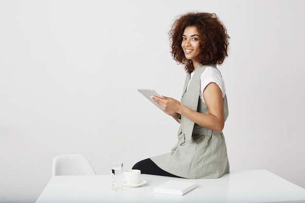 Muchacha africana hermosa que sonríe sosteniendo la tableta que se sienta en el lugar de trabajo. Copia espacio