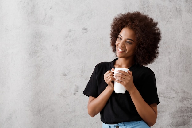 Muchacha africana hermosa que sostiene la taza sobre la pared ligera.