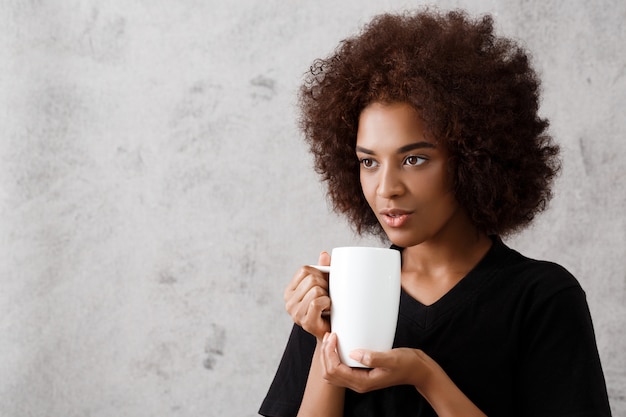 Muchacha africana hermosa que sostiene la taza sobre la pared ligera.