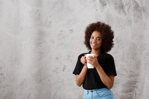 Muchacha africana hermosa que sostiene la taza sobre la pared ligera.