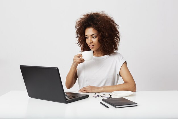 Muchacha africana joven que sostiene la taza que mira la computadora portátil que sonríe sobre la pared blanca.