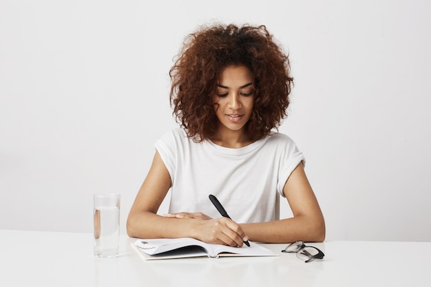 Muchacha africana que piensa escribir en el cuaderno que sonríe sobre la pared blanca. Copia espacio