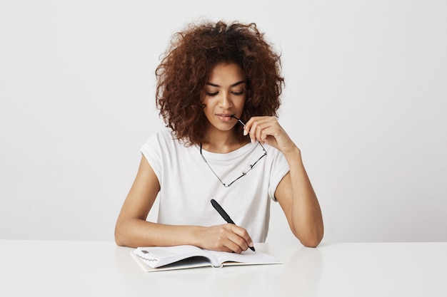 Muchacha africana que piensa escribir en el cuaderno que sonríe sobre la pared blanca. Copia espacio