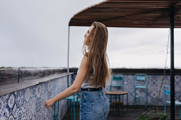 Muchacha agraciada con el pelo largo oscuro que presenta juguetonamente. Tiro al aire libre de mujer caucásica romántica viste falda de mezclilla.