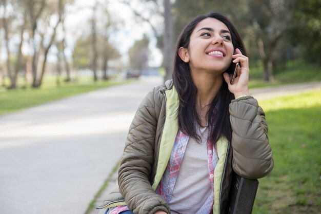 Muchacha alegre feliz del estudiante que habla en la célula en parque