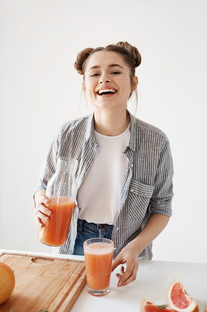 Muchacha alegre hermosa con los bollos que sonríen riendo sosteniendo el tarro de cristal con el smoothie sano de la desintoxicación del pomelo sobre la pared blanca.