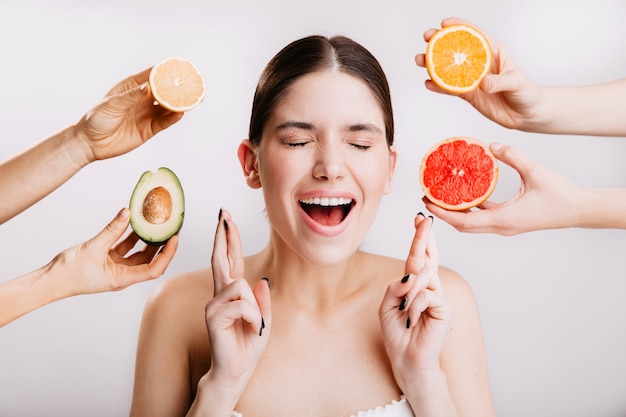 La muchacha alegre pide el deseo. Retrato de modelo sin maquillaje en pared blanca con frutas.