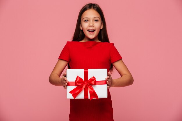 Muchacha alegre que muestra el regalo y la sonrisa aislada sobre rosa