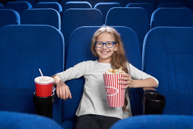 Muchacha alegre que sostiene el cubo de las palomitas de maíz, posando en cine.