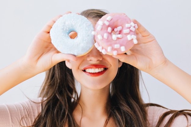 Muchacha alegre del retrato del primer que sostiene las rosquillas coloridas contra sus ojos. Atractiva mujer joven con pelo largo divirtiéndose con dulces deliciosos. Colores brillantes, concepto de dieta, dieta.