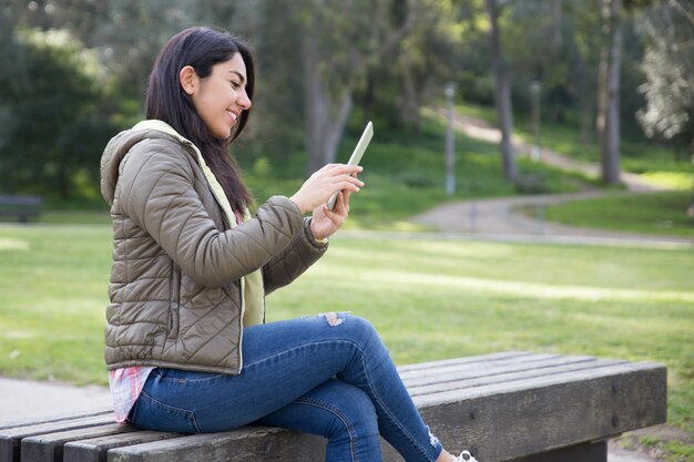 Muchacha alegre sonriente que disfruta de la conexión inalámbrica en parque de la ciudad