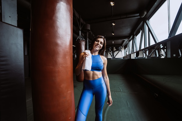 Muchacha apta sonriente que sostiene la toalla y que descansa en gimnasio cerca del saco de arena