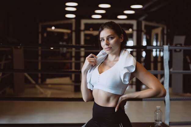 Muchacha apta sonriente que sostiene la toalla y que descansa en gimnasio.