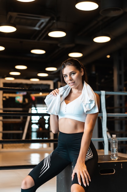 Muchacha apta sonriente que sostiene la toalla y que descansa en gimnasio.