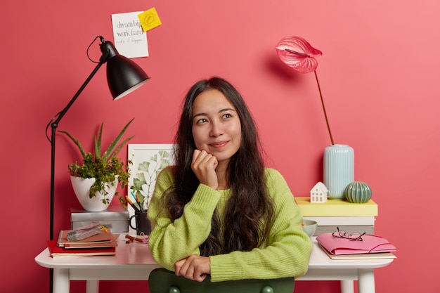 La muchacha asiática complacida se sienta contra el escritorio con todos los materiales necesarios para estudiar, sonríe y sueña despierto durante el descanso, posa en una silla contra la pared rosada brillante, notas adhesivas con inscripción.