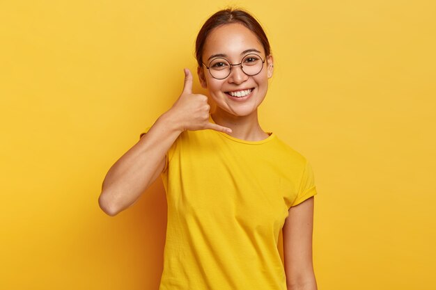 La muchacha asiática encantada sonriente muestra el gesto de llamarme, hace la señal de la mano del teléfono, tiene expresión feliz, piel sana, lleva gafas y ropa casual, aislada en la pared amarilla. Lenguaje corporal.