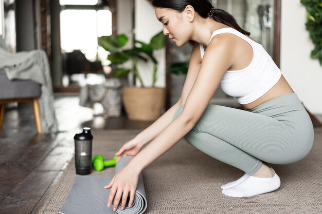 La muchacha asiática joven prepara la estera del piso para la meditación del yoga o el entrenamiento físico en el entrenamiento en casa en su casa ...