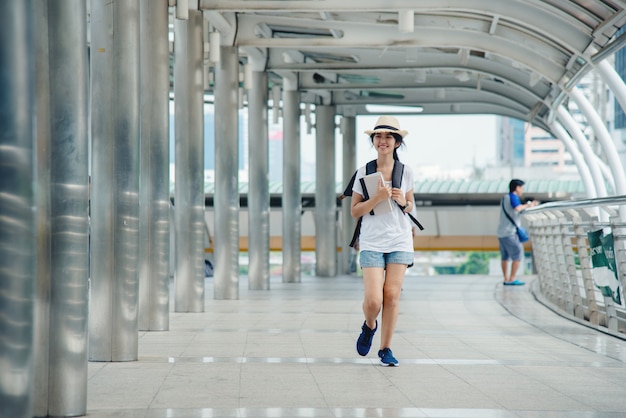 Muchacha asiática sonriente feliz del estudiante con la mochila en el fondo de la ciudad