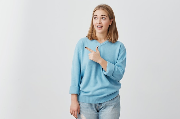 Muchacha asombrada con la boca abierta que mira con los ojos oscuros a un lado que señala con el dedo índice y que anuncia algo. Joven mujer posando con copia espacio para texto o promoción