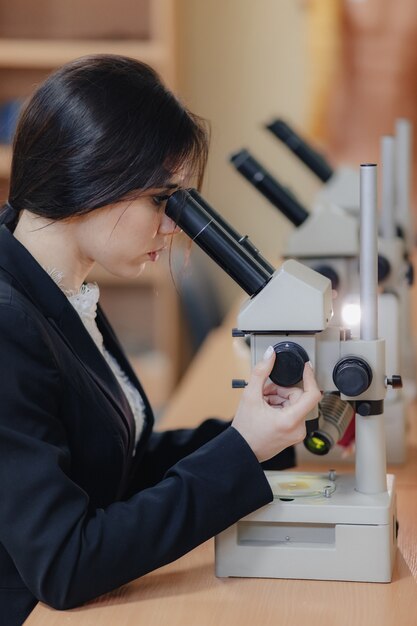 Muchacha atractiva emocional joven que se sienta en la tabla y que trabaja con un microscopio en una oficina o una audiencia moderna