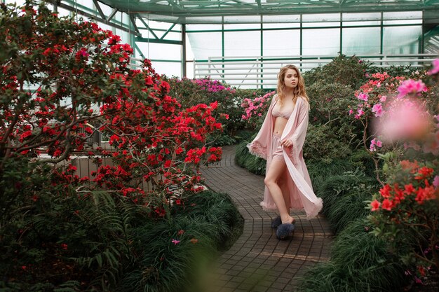 Muchacha atractiva hermosa que lleva la albornoz rosada y la ropa interior que se colocan en jardín de flores.