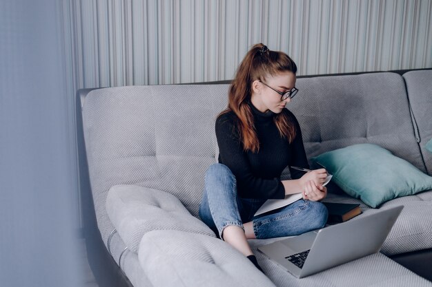 Muchacha atractiva joven en casa que trabaja con la computadora portátil en el sofá. Comodidad y comodidad en casa. oficina en casa y trabajo desde casa. empleo remoto en línea.