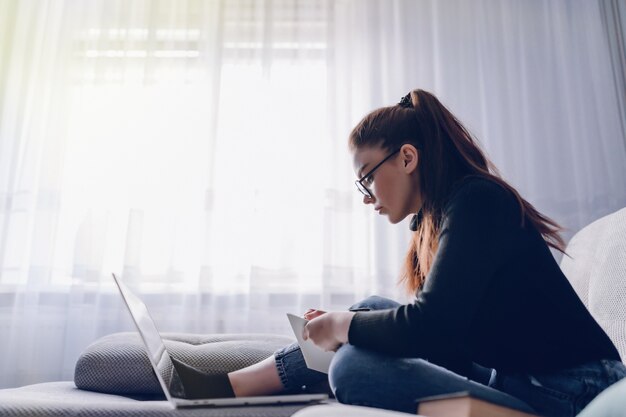 Muchacha atractiva joven en casa que trabaja con la computadora portátil en el sofá. Comodidad y comodidad en casa. oficina en casa y trabajo desde casa. empleo remoto en línea.